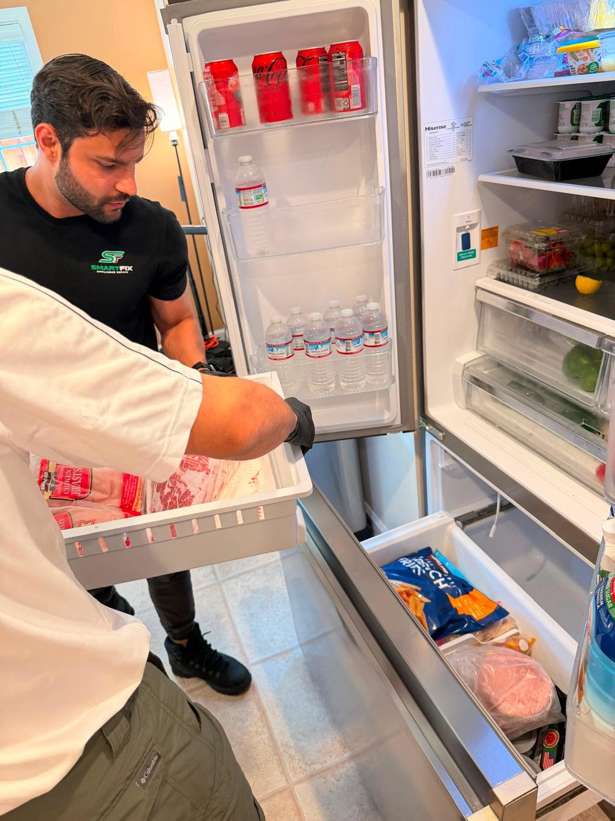 Technicians inspect an open refrigerator, one holding a freezer drawer filled with packaged meat.