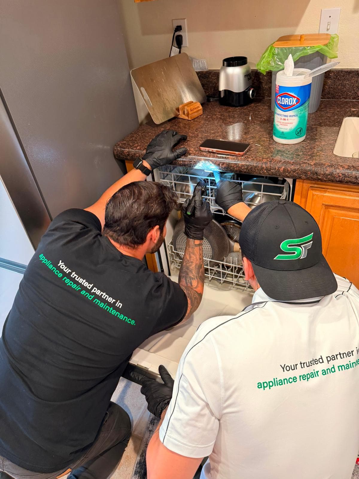 Two appliance repair technicians in branded shirts inspect a dishwasher in a home kitchen.