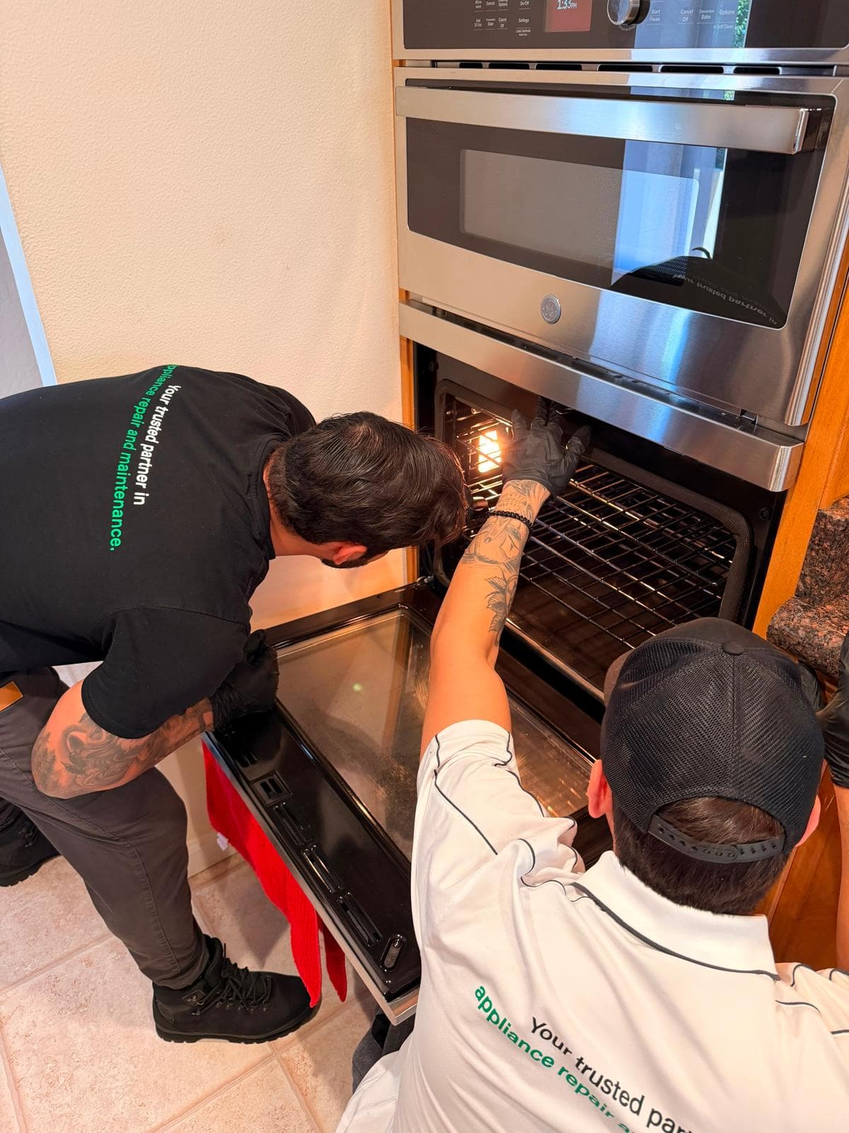 Two repair technicians in branded shirts inspect the interior of an open wall oven.