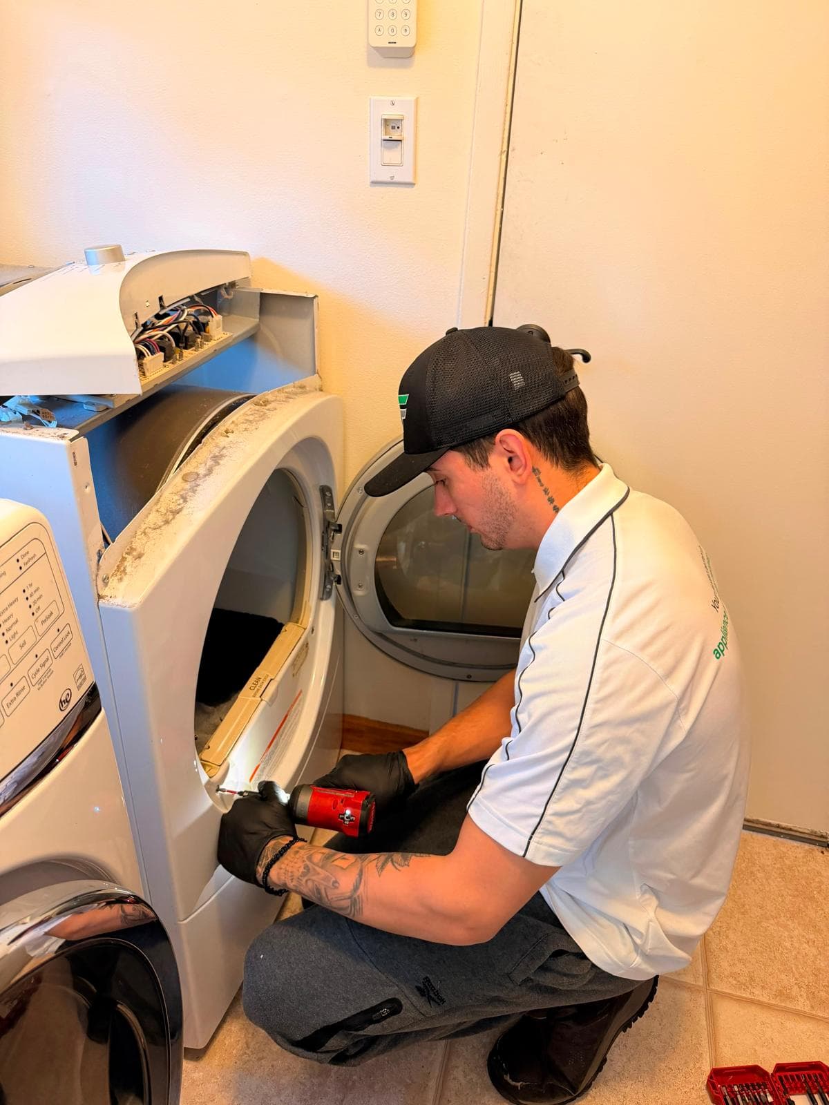 Technician in a white polo repairs a front-loading dryer using a red power drill.